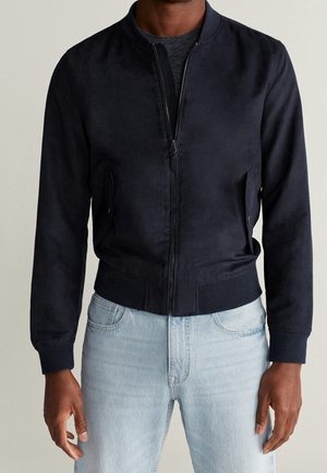 Homme portant une veste bomber noire ajustée avec une fermeture éclair avant, des poches à rabat, des poignets et un ourlet côtelés, associée à un jean bleu clair.