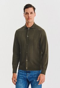 Oliwkowy kardigan z przodem na guziki, panelami o prążkowanej fakturze i klasycznym kołnierzykiem, w połączeniu z dżinsami.