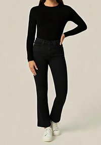 Femme portant un haut noir à manches longues, un pantalon évasé noir et des baskets blanches, debout avec une main sur la hanche.