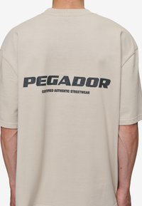 Valkoinen ylisuuri t-paita lyhyillä hihoilla, jossa on selässä iso musta teksti "PEGADOR" ja "CERTIFIED AUTHENTIC STREETWEAR".