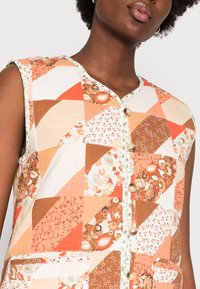 Chaleco de patchwork en tonos de naranja y marrón con patrones florales y geométricos. Presenta un cierre de botones y un diseño sin mangas, con bolsillos.