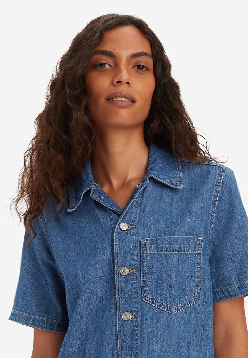 Chemise en denim à manches courtes, col classique et fermeture à boutons sur le devant. Dispose d'une poche poitrine et d'une texture délavée bleu.