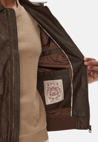 Interno di una giacca marrone imbottita con un patch con logo, tasca con zip e foderatura in satin liscio. La giacca ha polsini e colletto a coste.