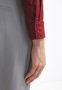 Chemise rouge à boutons avec une texture lisse, dotée d'un poignet ajusté à trois boutons rouges, associée à un pantalon en tissu gris.
