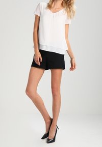 White short-sleeve blouse with a layered design, paired with black shorts. The outfit is complemented by black high-heeled shoes.