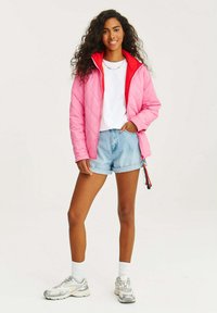 Prešívaná ružová bunda s červenými akcentmi, so zipsom a sťahovacou šnúrkou. Nosená cez biele tričko a spárovaná s ľahkými džínsovými šortkami a teniskami.
