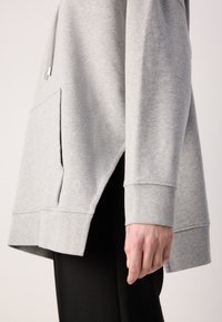 Sweatshirt gris clair à manches longues avec une poche avant et une fente latérale, porté avec un pantalon noir, mettant l'accent sur la zone du bras et de la taille.