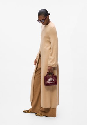 Beige long coat in soft fabric with wide sleeves, paired with brown trousers. Holds a small burgundy handbag with a logo. Leopard-print shoes.