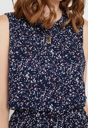 Mujer con parte superior azul marino sin mangas y parte inferior a juego con un patrón moteado en blanco, rojo y azul, y un collar con colgante rectangular dorado.
