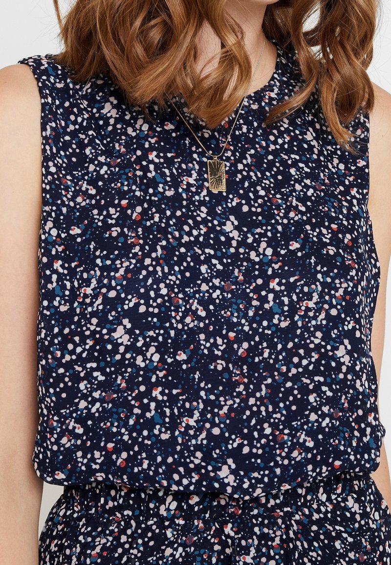 Femme portant un haut bleu marine sans manches et un bas assorti avec un motif tacheté blanc, rouge et bleu, ainsi qu'un collier pendentif rectangulaire en or.
