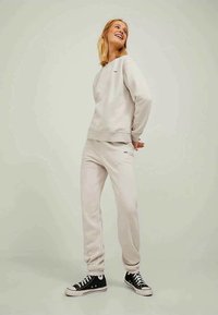 Light beige sweatshirt and matching sweatpants in soft fabric, featuring a logo on the chest and leg. Black sneakers complete the outfit.