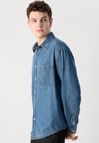 Camicia in denim con lavaggio medio, chiusura con bottoni frontale, colletto a punta, taschino sul petto e maniche lunghe con polsini abbottonati.
