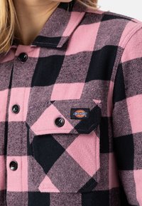 Camicia a quadri rosa e nera con bottoni in morbido flanella. Presenta una tasca sul petto con logo, bottoni neri e un colletto a risvolto.