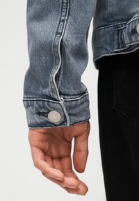 Χέρι χαλαρό στο πλευρό, που φοράει μανίκι ελαφρώς γαλάζιου τζιν μπουφάν με κουμπωμένο μανσέτα και μαύρο παντελόνι, με φόντο απλό.