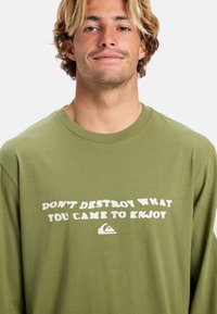 Langärmliges, olivgrünes Baumwoll-T-Shirt mit einem weißen, stilisierten Textdruck: "ZERSTÖRE NICHT, WAS DU GENIESSEN KONNTEST" und einem Logo.