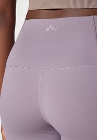 High-waisted lavender leggings made from stretchy fabric with flat seams and a small logo detail near the waistband. Smooth texture.