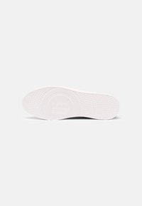 Semelle de chaussure blanche avec un motif de bande de roulement circulaire et en pointillés, incluant un logo en relief au centre, présenté sur un fond uni.