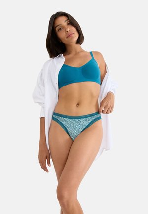 Woman wearing teal bra and patterned teal lace-trimmed underwear with white open shirt on light background.