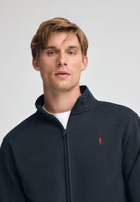 Giacca zip-up di un blu scuro, realizzata in tessuto liscio, con colletto alto e un piccolo logo rosso sul lato sinistro del petto. Sotto, una semplice maglietta bianca è visibile.