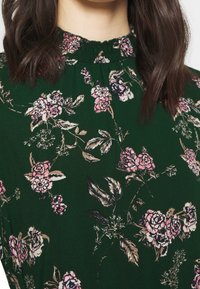 Grön textil med ett blommönster som har rosa rosor och gröna löv. Designen inkluderar en samlad halsringning och mjuk textur.