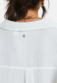 Witte linnen blouse met een opgestikte kraag, knoopsluiting aan de achterkant met logodetail en een subtiele gekreukelde textuur.