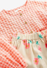 Blusa in gingham corallo con pattina a bottoni e maniche arricciate, abbinata a una gonna crema con motivo floreale rosa e fascia elastica in vita.