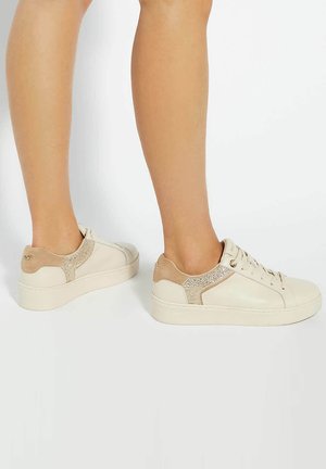 Baskets blanches avec des accents beiges, une tige texturée et une semelle plate. Comprend des lacets, un panneau pailleté à l'arrière et un design minimaliste.