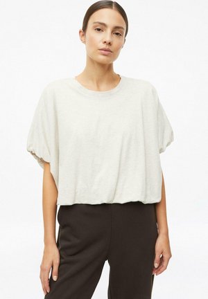 Vrouw met steil bruin haar die een losse lichtbeige top met korte mouwen en aangesloten donkerbruine broek draagt, poserend tegen een witte achtergrond.