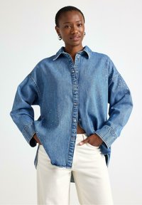 Camicia in jeans blu con colletto classico, bottoni frontali e decorata con piccoli dettagli in perline, abbinata a pantaloni avorio.