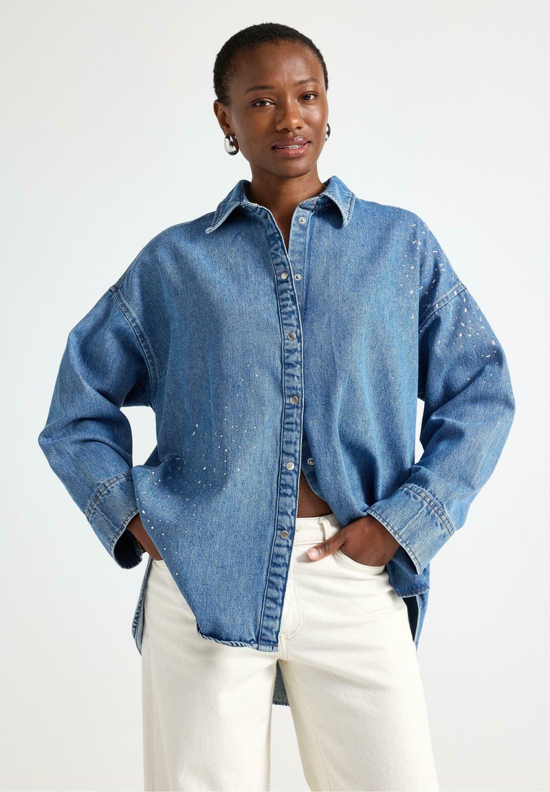 Camicia in jeans blu con colletto classico, bottoni frontali e decorata con piccoli dettagli in perline, abbinata a pantaloni avorio.