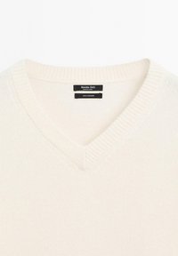 Halsnaht-V-Ausschnitt Pullover aus Kaschmir in Creme mit gerippter Hals- und Saumabschlüssen, glatter Textur und schwarzem Etikett, das Marke und Material angibt.