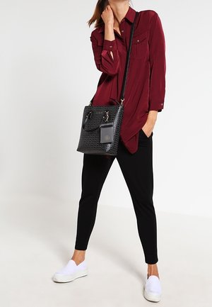 Femme portant un chemisier bordeaux, un pantalon noir et des chaussures blanches sans lacets, tenant un sac à main noir à motifs avec une petite pochette attachée.