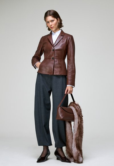 Femme portant un blazer en cuir marron avec un pantalon gris à jambes larges, tenant un sac à main marron avec une longue écharpe en fourrure sur un fond uni.