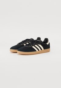 Chaussures de sport noires avec une tige texturée, trois rayures blanches et une semelle en caoutchouc gomme. Elles disposent d'un profil bas et d'un design à bout rond.