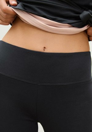 Mustat korkeavyötäröiset legginsit, joissa on sileä pinta ja leveä vyötärö, kerroksittain vaaleanpunaisen, hieman sisääntaitetun paidan alla.