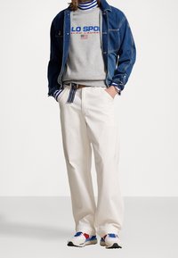 Džínová bunda přes šedou mikinu s modrým logem, bílé manšestráky a barevné tenisky s červenými, modrými a černými akcenty.