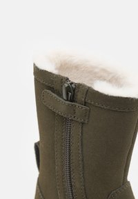 Olivengrønn semsket støvel med glidelås, myk hvit fôr og en ekstern løkke for enkel påkledning. Har en elegant og høy fasong.