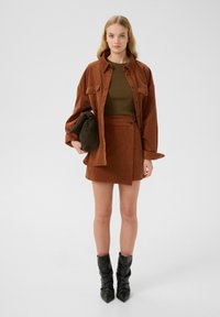 Set di camicia e gonna in corduroy ruggine con un top aderente verde oliva, abbinato a stivali alla caviglia neri e morbidi. La modella tiene una borsa verde.