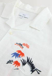 Chemise blanche à col avec un motif brodé composé de feuilles bleues, rouges et noires, ainsi qu'un oiseau noir. Tissu texturé subtil.
