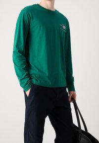 Zielona koszulka z długim rękawem z miękkiego materiału, z okrągłym dekoltem i haftowanymi zdobieniami na piersi. Noszona z ciemnymi spodniami i torbą.