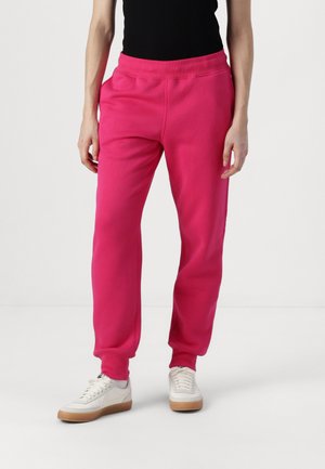 Rosa joggebukser laget av myk stoff, med elastisk midje og sidelommer, kombinert med hvite joggesko og en svart topp.