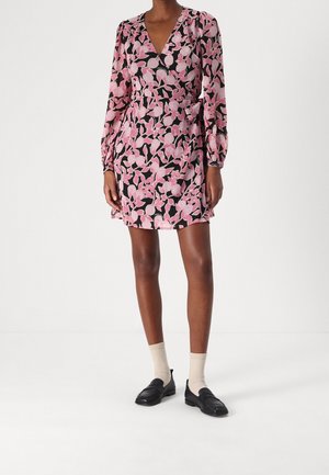 Vrouw die een roze-zwart bloemen wikkeljurk met lange mouwen draagt, beige sokken en zwarte loafers, staand tegen een witte achtergrond.