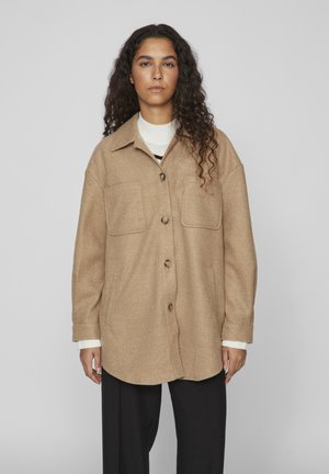 Beige Wollmischung-Übershirt mit spitzem Kragen, vorderem Knopfverschluss, zwei Brusttaschen und langen Ärmeln. Getragen über einem weißen Pullover.