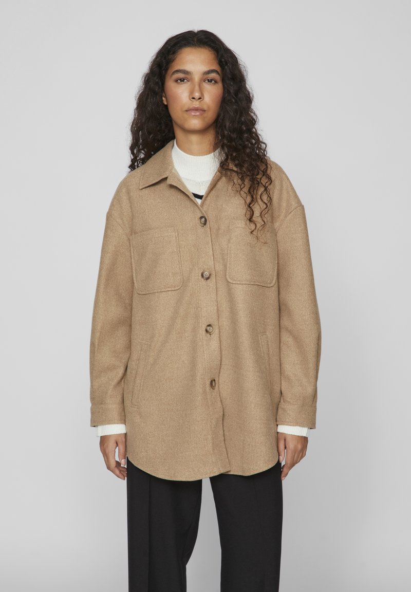 Beige Wollmischung-Übershirt mit spitzem Kragen, vorderem Knopfverschluss, zwei Brusttaschen und langen Ärmeln. Getragen über einem weißen Pullover.