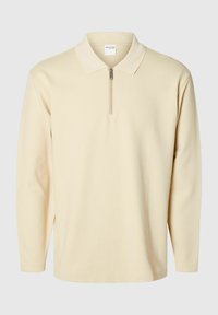 Beige poloko s dlouhým rukávem a zipem u krku, vyrobené z texturované látky, s klasickým límcem a postranními rozparky ve spodní části.