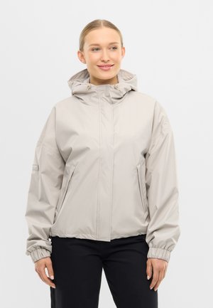 Nuori nainen pukeutuneena vaaleanbeigeen hupulliseen takkiin, jossa on joustavat hihansuut, ja mustiin housuihin, seisten yksinkertaisen taustan edessä.