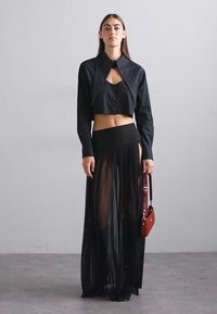 Μαύρο cropped πουκάμισο με μακριές μανσέτες και μυτερό γιακά, συνδυασμένο με μαύρη διάφανη maxi φούστα με πλάγια σχισμή. Το μοντέλο κρατά μια μικρή κόκκινη τσάντα.