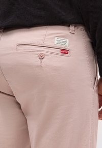 Κοντινό πλάνο στην πίσω τσέπη ροζ παντελονιού Levi's με κούμπωμα κουμπιού και δύο ορατές ετικέτες μάρκας στη δεξιά πλευρά.