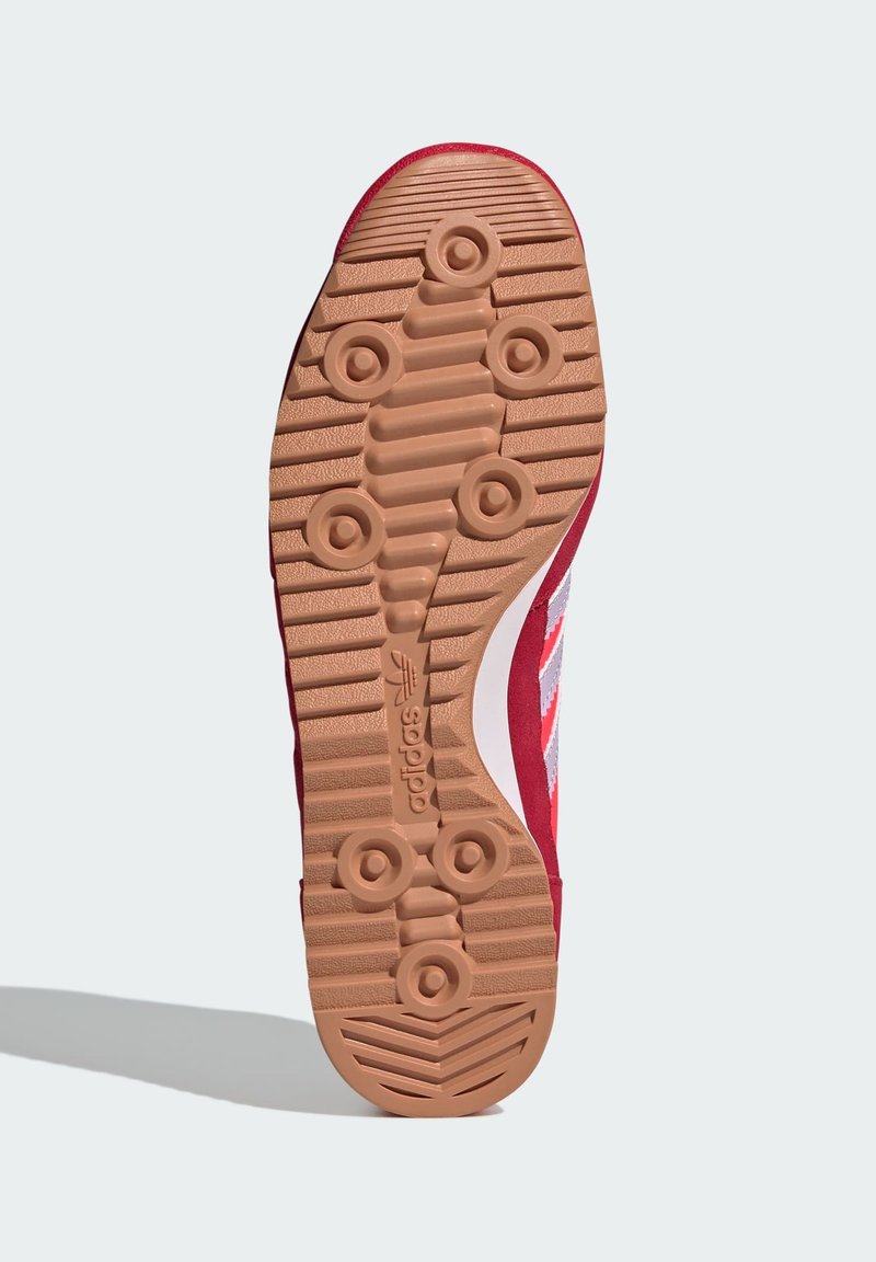 Zapatilla deportiva roja con suela de goma que presenta un patrón texturizado y tachuelas redondas. Rayas de acento blancas a lo largo del costado.