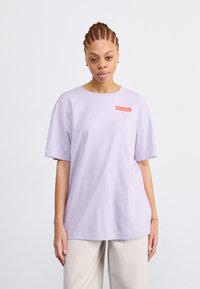 T-shirt oversized viola chiaro con scollatura rotonda e maniche corte. Presenta una toppa con logo rosso nella zona sinistra del petto.